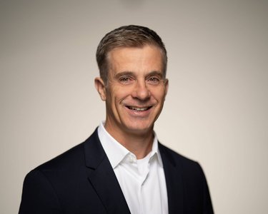 A smiling man with short, gray hair is wearing a dark suit and a white shirt. The background is neutral and bright.