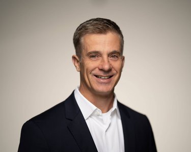 A smiling man with short, gray hair is wearing a dark suit and a white shirt. The background is neutral and bright.