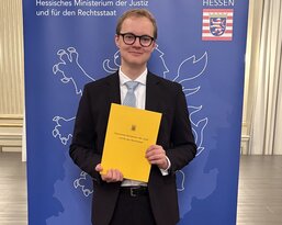 A young man in a suit is holding a yellow document and standing in front of a banner of the Hesse Ministry of Justice.
