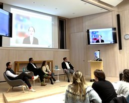 A discussion panel with four people sitting on stage while one person is connected via a screen.