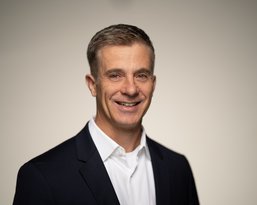 A man with short, gray hair is wearing a dark suit and a white shirt. He is smiling warmly at the camera.