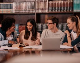 Four students are sitting at a table in a library, surrounded by books. They are discussing and working on their notes and a laptop.