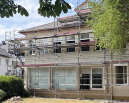 A building under scaffolding, surrounded by a garden with grass and bushes. The sky is partly cloudy.