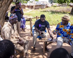 A group of men is sitting on benches under a tree, while two men in the middle are having a conversation.