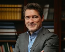 A man in a gray suit with a blue shirt stands smiling with his arms crossed in front of a bookshelf.