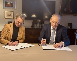 Two people are signing documents at a table indoors. A woman is wearing a beige blazer, and the man is in a suit.