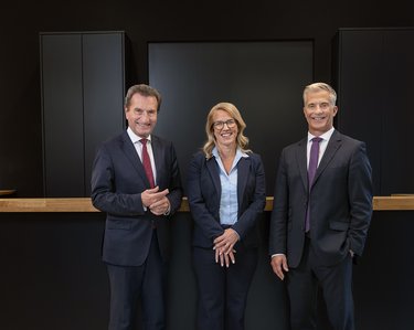 Three people in formal attire are standing side by side in a modern office building. The woman in the middle has short hair and is wearing glasses, while the two men on her left and right are smiling.
