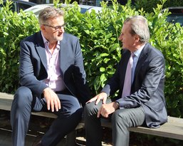 Two men are sitting on a bench, chatting, surrounded by green bushes. Both are dressed in formal attire.