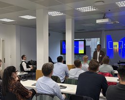 A speaker is presenting to a group of listeners in a modern seminar room. A presentation is visible on the wall.