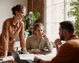 Three people in an office are discussing documents and notes while a laptop sits on the table. Plants are visible in the background.