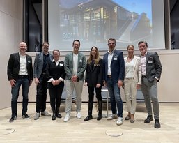 A group of eight people stands on a stage, smiling, dressed in formal attire, with a screen in the background.