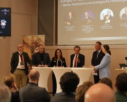 Discussion participants on a stage talking about European values and strategic approaches. Presentation slides are visible in the background.