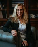 A blonde woman with glasses is sitting on a sofa, wearing a black blazer and a white shirt, and smiling warmly.