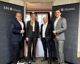Four people stand in front of two banners that read 'EBS Alumni.' They are dressed in formal attire and are posing cheerfully.