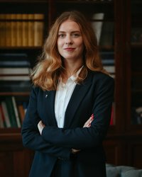 A woman with long, curly hair stands with her arms crossed in a suit in front of a bookshelf.