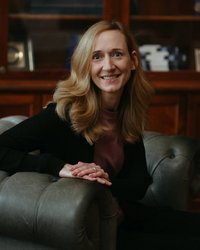 A woman with long, curly hair is sitting in an armchair and smiling. She is wearing a black sweater and a purple blouse.