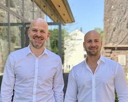 Two men in white shirts are smiling in front of a modern building with a historic tower in the background.