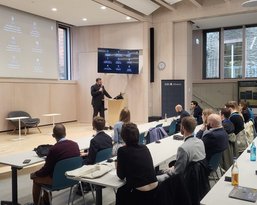 A speaker stands at a podium and addresses an audience in a modern event space with large screens.