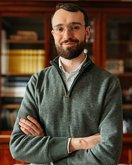 A man with glasses and a beard stands with his arms crossed in a gray sweater in front of a bookshelf.