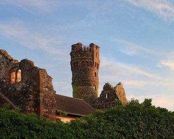Ruins of a stone castle with a tall tower, surrounded by lush greenery and a low wall, under a clear sky at sunset.