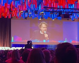 A man is speaking on a stage while the audience sits in the foreground. Colorful light installations hang from the ceiling.