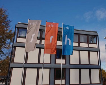 Three flags bearing the letters 's', 'r', and 'h' stand in front of a modern building, while another state-of-the-art structure is visible in the background. The sky is partly cloudy.