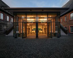 Modern glass doors lead into a brightly lit interior, surrounded by historic buildings with red window frames. The floor is made of cobblestones that encircle the entrance area.