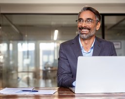 A smiling man with glasses sits at a table in front of a laptop, surrounded by office supplies in a modern office.
